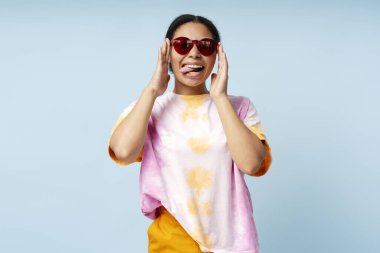 Güzel gülümseyen Afro-Amerikan kadın portresi. Modaya uygun kravat boyası tişört ve mavi arka planda izole güneş gözlüğü takıyor. Gülümseyen moda modeli stüdyoda poz verirken eğleniyor. 