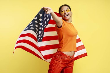 Güzel gülümseyen Afro-Amerikan kadın sarı arka planda Amerikan bayrağı taşıyor. Genç mutlu vatanseverlerin Bağımsızlık Günü kutlamalarının portresi başka yöne bakıyor. 4 Temmuz konsepti