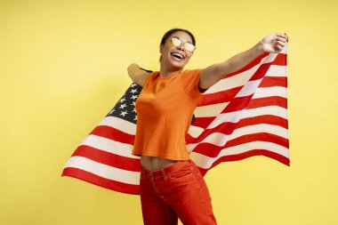 Güzel gülümseyen Afro-Amerikan kadın sarı arka planda Amerikan bayrağı taşıyor. Genç mutlu vatanseverlerin Bağımsızlık Günü kutlamalarının portresi başka yöne bakıyor. 4 Temmuz konsepti 