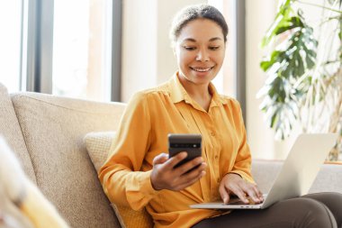 Modern gülümseyen Afro-Amerikan iş kadını, cep telefonu tutan serbest çalışan, bilgisayarını evde çalışan. Akıllı telefonlu şık bir metin yazarı kafede oturuyor. Başarılı bir iş.