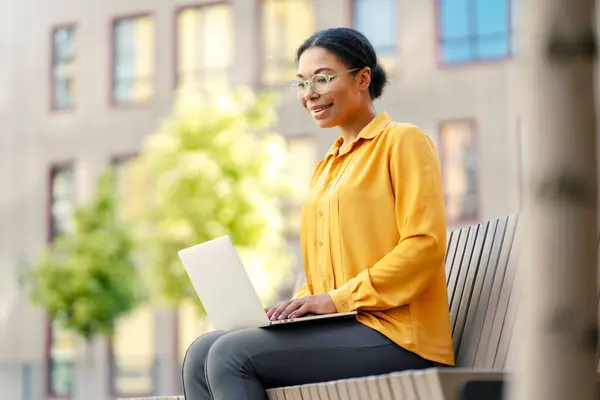 Beautiful smiling African American woman, worker holding laptop computer, having video call sitting on bench outdoors. Modern technology, video conference, online lesson concept