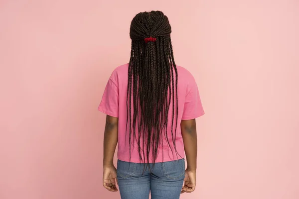 Back view portrait of authentic African girl with long hair, dreadlocks, pigtails, wearing pink t shirt and jeans isolated on pink background. Hair care concept