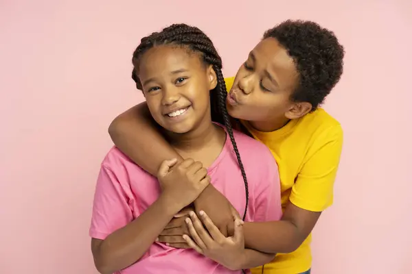 Portrait of smiling African American brother and sister hugging, kissing isolated on pink background
