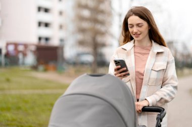 Neşeli anne bebek arabasını bebekle itiyor, telefon kullanıyor, sokakta mesajlaşıyor, parkta mesajlaşıyor. Gülümseyen kadın yürüyüşü, sağlıklı yaşam tarzı konsepti