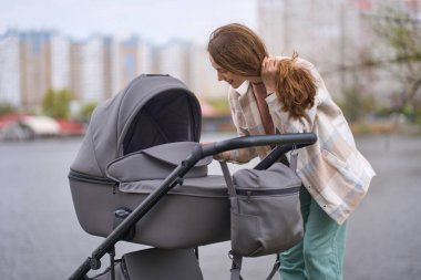 Genç mutlu anne uyuyan bebeğiyle bebek arabasına bakıyor, ılık bir bahar gününde şehir parkında yürürken tatlı tatlı gülümsüyor. Beyaz kadın anne doğum izninin tadını çıkarıyor. İnsanlar ve Yaşam Tarzı