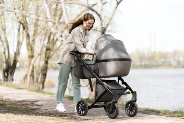 Modern anne, bebek arabasıyla bebek arabasını iterken ona dışarıda bakıyor. Mutlu annelik, bakım, ebeveynlik, sağlıklı yaşam tarzı.