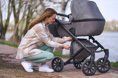 Genç anne, beyaz saçlı güzel bir kadın, günlük giysiler içinde, yeni doğmuş çocuğuyla nehir kenarında parkta yürürken bir bebek arabasının bagajını açıyor..