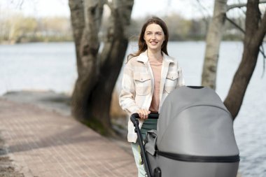 Küçük bir çocukla bebek arabasını iten mutlu genç anne, doğada, açık havada, göl kenarında yürüyor. Mutlu kadın, güneşli gün