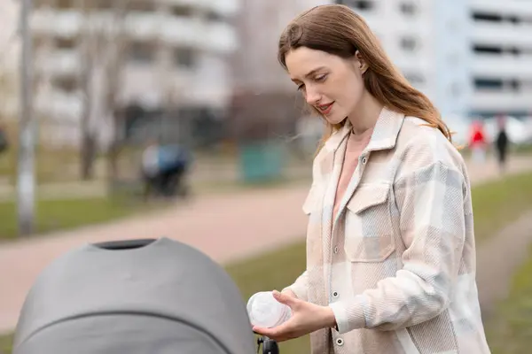 現代の若い母親は 馬車で新生児と屋外を歩き 赤ん坊のボトルを手に持っていました 子供と公園でカジュアルな服で幸せな女性 — ストック写真