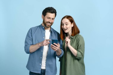 Kafkasyalı gülümseyen yetişkin çift cep telefonu kullanıyor, haber yayını çekiyor, sosyal medya içeriğini kontrol ediyor, çevrimiçi mesajlar atıyor, yeni cep telefonu uygulamalarını kontrol ediyor. İzole edilmiş mavi renk stüdyo arka planı