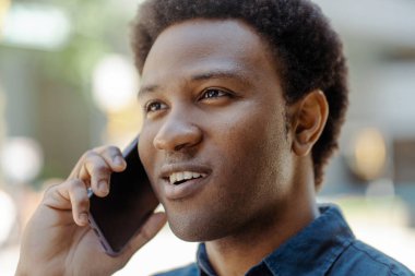 Gülümseyen yakışıklı Afro-Amerikan iş adamının portresi cep telefonuyla konuşuyor, telefona cevap veriyor ve şehir sokağına bakıyor. Başarılı bir iş.