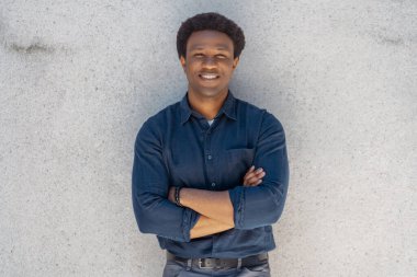 Kollarını kavuşturmuş gri duvarın yanında duran gülümseyen yakışıklı Afrikalı adamın portresi. Çekici bir erkek, günlük giysiler içinde fotoğraf için poz veriyor.