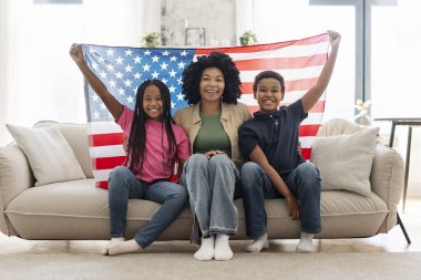 Afrika kökenli Amerikalı kadın ve çocukların portresi, elinde Amerikan bayrağı, Amerika 'nın ulusal sembolü, oturma odasında, evinde, kameraya bakarak kanepede oturuyor. Vatanseverlik kavramı