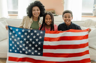Çekici Afro-Amerikan kadın ve çocukların portresi, elinde Amerikan bayrağı, Amerika 'nın ulusal sembolü, oturma odasında kanepede oturuyor, evde kameraya bakıyor. Vatanseverlik kavramı