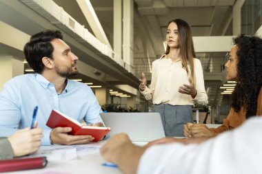 Kendine güvenen bir grup iş adamı, çekici genç kadın konuşuyor, beyin fırtınası yapıyor, modern ofiste planlama stratejisi uyguluyor. İş kadını bir şeyi açıklıyor. Takım çalışması, toplantı, seminer, başarılı iş