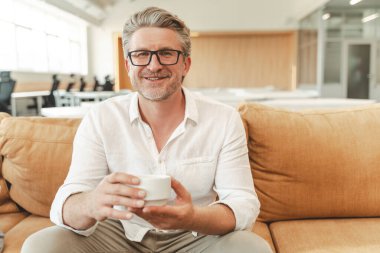 Gülümseyen, kendine güvenen gri saçlı işadamının portresi, modern ofiste ekonomi sınıfında oturan kameraya bakan bir işçi. Günaydın, kahve molası. 