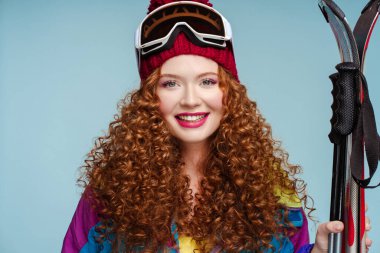 Kırmızı dudaklı, kayak yapan, kar gözlüklü, tulumlu, mavi arka planda izole edilmiş kameraya bakan çekici kıvırcık kadın. Kış sporları