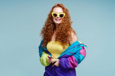 Kış tulumlu kızıl saçlı güzel bir kadın güneş gözlüğü takıyor ve mavi arka planda izole edilmiş kameraya bakıyor. Moda konsepti