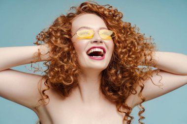 Mavi arka planda izole edilmiş, güneş gözlüğü takan heyecanlı, kıvırcık saçlı kadının portresi. Moda konsepti, reklam