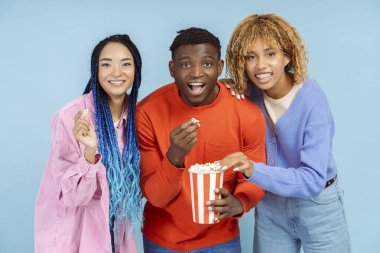 Bir grup gülümseyen stil sahibi Afrikalı Amerikalı arkadaşlar patlamış mısır yiyor ve mavi arka planda izole bir şekilde film izliyorlar.