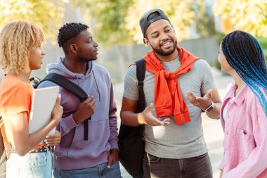 Bir grup gülümseyen Afro-Amerikan arkadaş renkli günlük giysiler içinde sokakta üniversite kampüsünün yakınında konuşuyorlar. Dışarıda mutlu öğrenciler. Eğitim, iletişim, buluşma kavramı