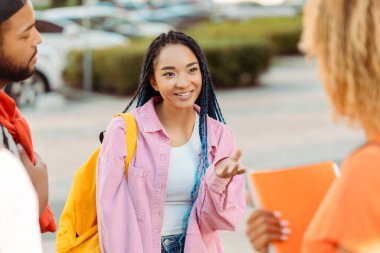 Güzel gülümseyen Afro-Amerikan bir kadının portresi. Üniversite kampüsündeki arkadaşlarıyla sarı sırt çantalı iletişim kuran zeki bir öğrenci. Eğitim kavramı