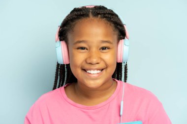 Kulaklıkla müzik dinleyen gülümseyen Afrikalı bir kızın portresi, günlük pembe tişört giyiyor, mavi arka planda tek başına duruyor, kameraya bakıyor, kapatıyor.