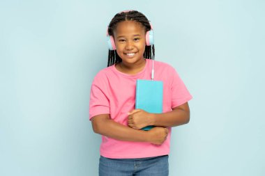 Mavi arka planda izole edilmiş kulaklıkla müzik dinleyen gülümseyen Afrikalı Amerikalı kız portresi