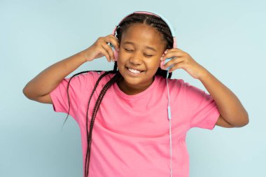 Gözleri kapalı, mavi arka planda izole edilmiş kulaklıkla müzik dinleyen, gülümseyen Afro-Amerikan bir kızın portresi. Sevimli gülümseyen küçük bayan eğleniyor