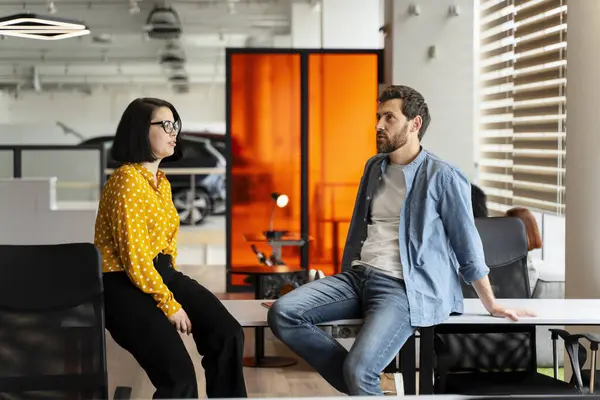 stock image Young bearded Hispanic man in casual denim sitting relaxed on the office table and talking with his colleague in friendly office atmosphere. Team. People. Coworkers. Business and communication