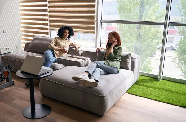View from above. Two diverse millennial women, colleagues, business partners talking about business project in friendly relax atmosphere, sitting together on a sofa in modern corporate office interior