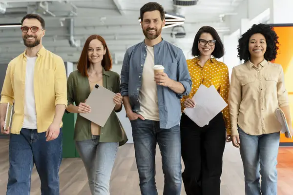 Diverse group of successful office employees, sales force managers, in casual wear, walking in a modern office interior, smiling looking at camera. Team. People. Coworkers. Business and communication