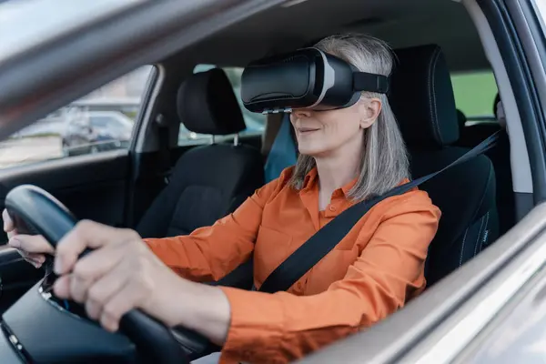 Confident portrait of a gray-haired Caucasian senior woman 60 years old, wearing virtual reality glasses, holding hands on the steering wheel, driving a car with a fastened seat belt. Road insurance.