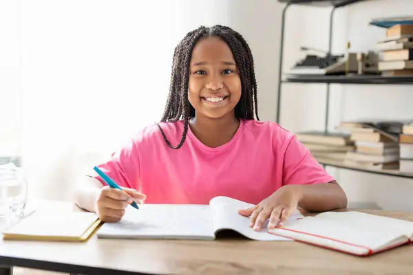 テーブルに座ったスタイリッシュなピンクのTシャツを着た魅力的なアフリカ系アメリカ人の少女の肖像画 ペンを保持し メモを取る 言語を学ぶ カメラを見る 学校のコンセプトに戻る — ストック写真