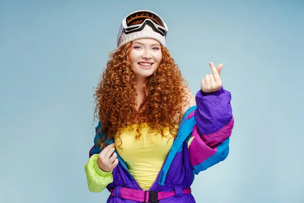 一个有着卷曲头发的微笑的女人的画像 用手指表示她的心 K流行文化 蓝色背景隔离 年轻的现代女性 戴着滑雪护目镜 看着相机 — 图库照片