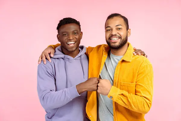 stock image Portrait of smiling positive African American male friends in casual colorful outfit giving fist bump, hugging, looking at camera isolated on pink background. Friendship concept