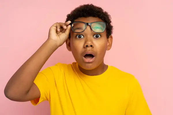 衝撃を受けた魅力的なアフリカ系アメリカ人の少年が眼鏡をかけ ピンクの背景に隔離されたカメラを見ているカジュアルな黄色のTシャツ ハンサムな学校の男性は驚きました — ストック写真