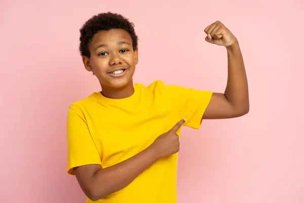 一个面带微笑 强壮的非洲裔美国青少年的画像 用手指指指二头肌 身穿休闲T恤 背景为粉色 迷人的儿童姿势 在工作室里看着相机 — 图库照片