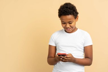Beyaz tişörtlü, gülümseyen Afro-Amerikan çocuğun portresi cep telefonu, mesaj, sipariş, online alışveriş bej arka planda izole edilmiş, kopyalama alanı. Sosyal medya kavramı