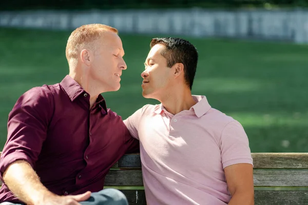 Portrait of attractive two gay men hugging and kissing in park, sitting in park on bench. Date, love concept
