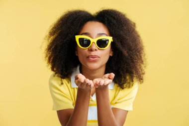 Sarı arka planda izole edilmiş, havalı sarı gözlük takan, gülümseyen, kıvırcık saçlı, güzel Afro-Amerikan kadının portresi. Moda modeli konsepti