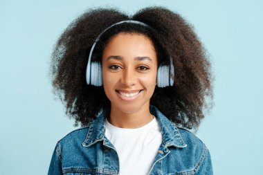 Kıvırcık saçlı, şık kot ceket giyen, kulaklıkla müzik dinleyen, mavi arka planda izole edilmiş kameraya bakan çekici Afro-Amerikan kadın portresi. Teknoloji konsepti