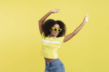 Sarı arka planda izole edilmiş şık sarı güneş gözlüğü takan kıvırcık saçlı, gülümseyen Afro-Amerikan genç kadın portresi. Yaz konsepti