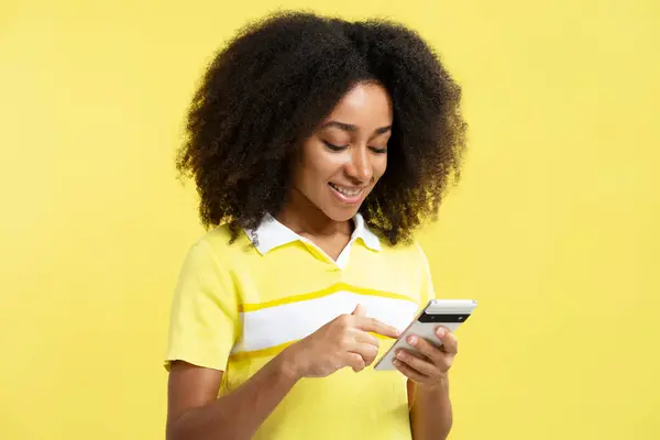 Beautiful smiling African American woman holding smartphone, using mobile app shopping online isolated on yellow background. Happy curly haired hipster female chatting. Technology concept