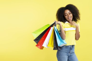 Sarı arka planda izole edilmiş renkli alışveriş torbaları taşıyan mutlu kıvırcık saçlı Afrikalı Amerikalı kadın. Mağaza, alışveriş, satış, Kara Cuma konsepti