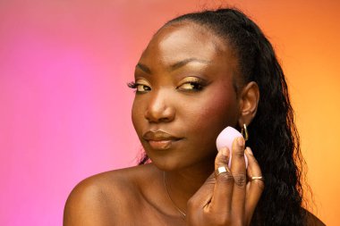 Güzel Afro-Amerikalı bir kadın, renkli arka planda tek başına dururken güzellik blenderi süngeri kullanıyor. Cilt bakımı ve güzellik kavramı