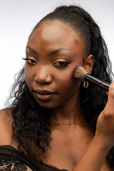 化粧ブラシを使った若いアフリカ系アメリカ人女性は 白い背景に隔離されたカメラを見ながら装飾的なメイクを施しています 化粧品コンセプト — ストック写真