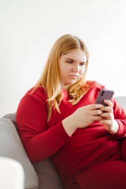 Kırmızı elbiseli, cep telefonlu, çekici genç bir kadın oturma odasındaki kanepede oturup mesajlaşarak sohbet ediyor. Teknoloji konsepti