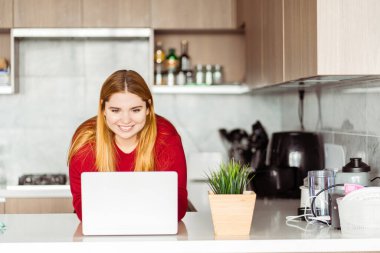Güzel, gülümseyen bir kadın ayakta, dizüstü bilgisayar kullanıyor, modern bir evde, mutfakta çalışıyor. Uzak görev kavramı
