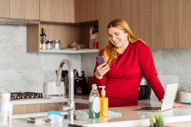 Güzel, iri yarı, kırmızı günlük kıyafetler giyen, cep telefonu tutan, e-postaları kontrol eden, dizüstü bilgisayarını kullanan, evde, mutfakta çalışan bir kadının portresi. Uzak görev konsepti